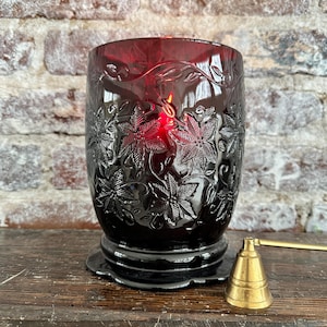 May include: A lit, dark red glass candle holder with a floral design. The candle holder sits on a black base and has a brass candle snuffer next to it. The candle flame is visible inside the holder.