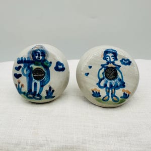 Puede incluir: Dos pomos de cerámica para armarios con diseños pintados a mano en azul y blanco. Un pomo presenta una figura tocando la flauta, el otro una figura con un vestido. Ambos tienen tornillos negros y detalles florales.