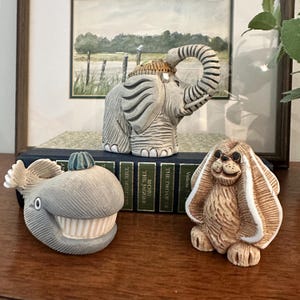May include: Three ceramic animal figurines: a gray elephant with a raised trunk, a gray whale with a prominent toothy grin and a small blue fin, and a tan long-eared dog with dark eyes. The figurines are arranged on a dark green book spine.