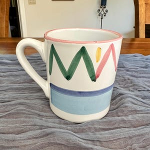 May include: A white ceramic mug featuring a hand-painted design with a pink rim, green zigzag lines, a small yellow dot, and a horizontal band consisting of a dark blue stripe above a wider light blue stripe, complete with a curved side handle.