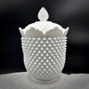 May include: A white hobnail glass jar with a lid. The jar has a rounded body covered in small, raised bumps. The lid features a scalloped edge and a teardrop-shaped finial. The jar sits on a white surface against a black background.