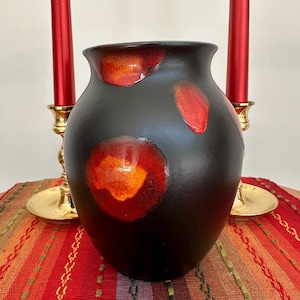 Könnte beinhalten: Eine schwarze Keramikvase mit roten und orangefarbenen kreisförmigen Designs. Die Vase hat eine abgerundete Form und einen leicht ausgestellten Rand. Zwei rote Kerzen in goldenen Kerzenhaltern flankieren die Vase.