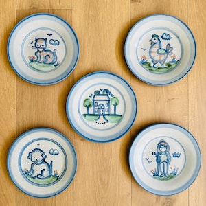 Puede incluir: Conjunto de cinco platos de cerámica con bordes azules e ilustraciones pintadas a mano. Cada plato presenta un diseño diferente: un gato, un gallo, una casa, un perro y una persona. Los platos tienen una estética vintage y de arte popular.