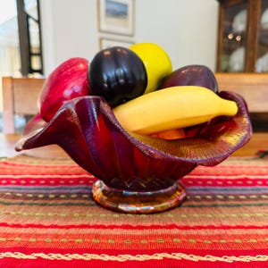 Puede incluir: Un frutero decorativo de cristal rojo iridiscente lleno de fruta artificial. El cuenco contiene una manzana roja, ciruelas moradas oscuras, un plátano amarillo y una manzana amarilla. El cuenco se encuentra sobre un camino de mesa a rayas rojas y naranjas.