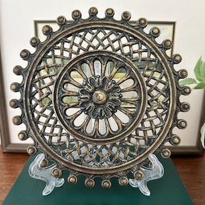 Könnte beinhalten: Eine kreisförmige, dekorative Wandplatte mit dunkelbronzefarbenem Finish und goldenen Akzenten. Das Design umfasst ein zentrales Blumenmotiv, umgeben von einem Gittermuster, und einen gewellten Außenrand, der mit kleinen erhabenen Kugeln verziert ist.
