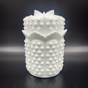 Puede incluir: Un tarro blanco estilo hobnail con tapa. El tarro tiene una base redonda y un borde festoneado. La tapa está rematada con un diseño en forma de lazo. El tarro es de vidrio y tiene un acabado brillante.
