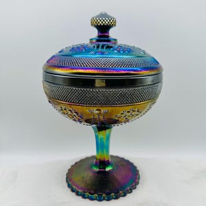 Könnte beinhalten: Eine irisierende Bonbonniere aus Glas mit Deckel. Die Schale hat eine runde Schalenform mit einer strukturierten, gemusterten Oberfläche. Die Farben wechseln von Blau über Gold zu Lila. Die Schale steht auf einem Sockel.