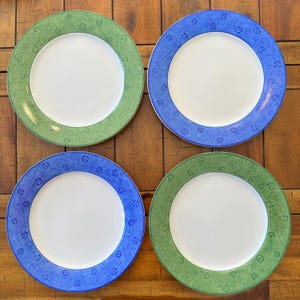 Puede incluir: Cuatro platos de cerámica blanca con bordes decorativos. Dos platos tienen un borde verde con un diseño de remolino, y dos tienen un borde azul con un patrón cuadrado. Los platos están dispuestos sobre una superficie de madera.