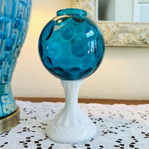 Może przedstawiać: Turkusowy szklany wazon w kształcie kuli z białą podstawą. Kula ma wzór tekstury z okrągłymi wgłębieniami. Podstawa jest biała, z żłobionym wzorem. Wazon to dekoracyjny element wystroju domu.