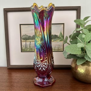 Peut inclure: Un grand vase en verre irisé présentant une couleur de base rouge profond avec une finition arc-en-ciel chatoyante. Le vase possède un bord évasé et festonné, un col fin et un corps texturé à motifs de diamants sur une base hexagonale.