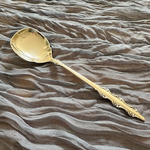 Puede incluir: Una cuchara de servir en tono dorado pulido con un cuenco ancho y redondeado y un mango delgado. El mango está decorado con un patrón floral en relieve a lo largo de su longitud, terminando en una punta. La superficie es metálica y lisa.