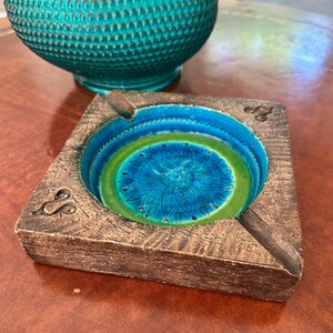 May include: A square ashtray with a textured brown exterior and a circular interior basin featuring a vibrant blue and green gradient glaze. The center of the basin displays a detailed dark blue floral pattern, and the corners feature carved symbols.