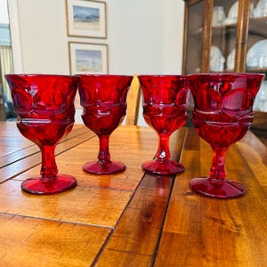 Puede incluir: Cuatro copas de vino vintage de color rojo rubí con un patrón texturizado. Las copas tienen una forma de cáliz clásica con una base redonda y un tallo largo. Las copas están dispuestas sobre una mesa de madera.
