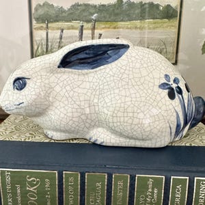 May include: A ceramic rabbit figurine featuring a white base with a dark blue crackle glaze finish. The rabbit has dark blue painted ears, eyes, and a floral motif on its side. It is sculpted in a resting position with its ears laid back.