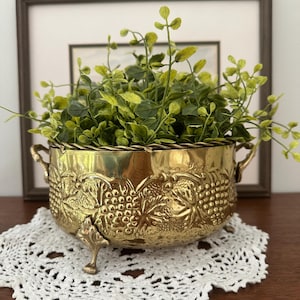 Puede incluir: Una maceta de latón pulido con cuerpo redondeado que presenta motivos en relieve de uvas y hojas. El recipiente incluye dos asas laterales, tres patas ornamentadas y un borde de cuerda trenzada, conteniendo una planta artificial verde.