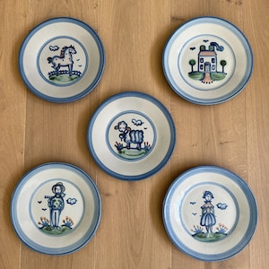 Puede incluir: Cinco platos de cerámica con diseños pintados a mano. Cada plato presenta un centro blanco con un borde azul y una ilustración única: un caballo, una casa, una oveja, una persona con un rastrillo y una persona con un vestido.