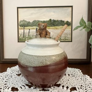 May include: A ceramic honey pot with a round body featuring a deep red base that transitions into a speckled light gray middle and a white rim. The lid is topped with a small brown bear figurine and includes a wooden honey dipper inserted inside.