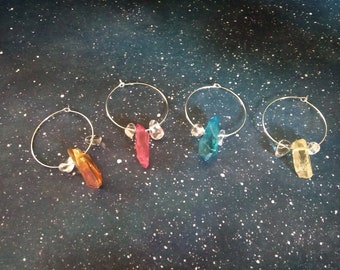 May include: Four silver hoop earrings, each adorned with a different colored crystal: orange, red, teal, and yellow. Clear crystal accents are also present. The earrings are displayed against a dark blue background with white speckles.