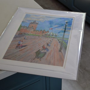 May include: A framed watercolor painting of a boardwalk scene with a large building in the background. The boardwalk is filled with people and benches. The sky is blue with white clouds. The painting is in a white frame with a white mat.