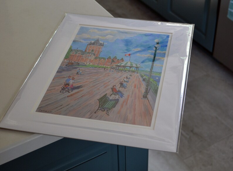 May include: A framed watercolor painting of a boardwalk scene with a large building in the background. The boardwalk is filled with people and benches. The sky is blue with white clouds. The painting is in a white frame with a white mat.