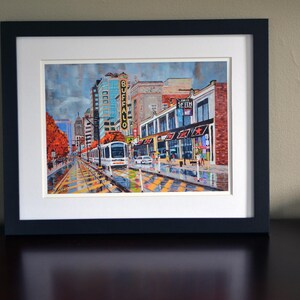 May include: A framed painting of a city street scene with a light rail train in the foreground. The train is white with a yellow stripe and the words "BUFFALO" in yellow letters on the side. The buildings in the background are brick and have storefronts with awnings. The street is wet and there are people walking on the sidewalk.