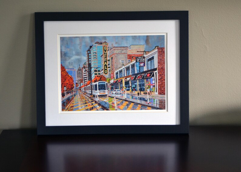 May include: A framed painting of a city street scene with a light rail train in the foreground. The train is white with a yellow stripe and the words "BUFFALO" in yellow letters on the side. The buildings in the background are brick and have storefronts with awnings. The street is wet and there are people walking on the sidewalk.