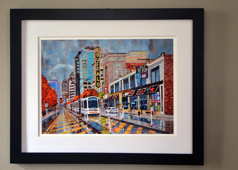 May include: A painting of a city street scene with a streetcar in the foreground. The streetcar is white with red and blue accents. The buildings in the background are brick and have signs that say "Buffalo" and "Shea's Smith Music Hall".