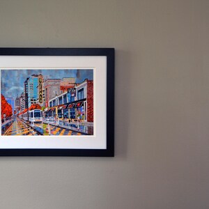 May include: A framed painting of a city street scene with a streetcar in the foreground. The buildings are brick and have storefronts. The streetcar is yellow and has the word "Buffalo" on the side. The painting is in a black frame.