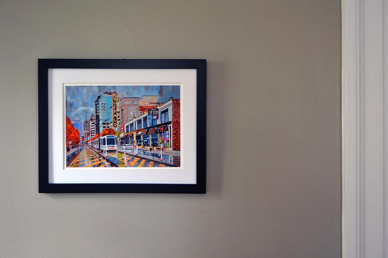 May include: A framed painting of a city street scene with a streetcar in the foreground. The buildings are brick and have storefronts. The streetcar is yellow and has the word "Buffalo" on the side. The painting is in a black frame.