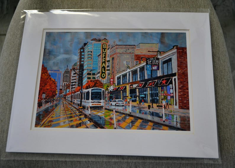 May include: A painting of a city street scene with a streetcar in the foreground. The streetcar is white with a red stripe and is traveling down a street lined with buildings. The buildings are brick and have storefronts. There are trees on either side of the street and people walking on the sidewalk. The sky is cloudy and there is a sign that says "Buffalo" on one of the buildings.