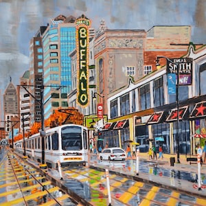 May include: A white streetcar travels down a city street with tall buildings and a sign that reads "Shea's Buffalo" in the background. The street is wet from rain and there are yellow and black markings on the street.