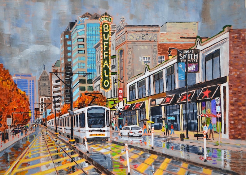 May include: A white streetcar travels down a city street with tall buildings and a sign that reads "Shea's Buffalo" in the background. The street is wet from rain and there are yellow and black markings on the street.