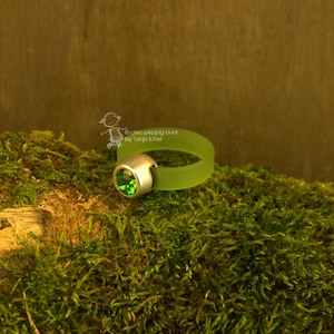 Könnte beinhalten: Ein grüner Ring mit silberner Fassung und einem großen, runden, grünen Edelstein. Das Ringband ist transparent grün. Der Ring wird auf grünem Moos präsentiert.
