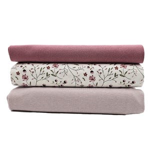 Könnte beinhalten: Drei Stoffrollen übereinander gestapelt. Die oberste Rolle ist einfarbig mauve. Die mittlere Rolle ist weiß mit einem Blumenmuster in Mauve und Grün. Die untere Rolle ist mauvefarben mit dünnen, horizontalen Streifen.