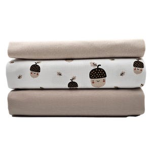 Könnte beinhalten: Drei Schichten gefalteten Stoffes. Die oberste Schicht ist ein gerippter, hellbeige Stoff. Die mittlere Schicht ist weiß mit einem sich wiederholenden Muster aus lächelnden Eicheln in Braun und Beige. Die untere Schicht ist einfarbiger, hellbeige Stoff. Die Stoffe sind ordentlich gestapelt.