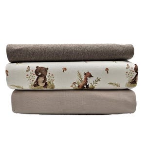Könnte beinhalten: Drei Stoffstapel. Die oberste und unterste Schicht sind einfarbig braun und beige. Die mittlere Schicht ist hellbeige mit einem sich wiederholenden Muster aus Bär, Fuchs, Pilzen und Schmetterlingen in einer Waldszene.