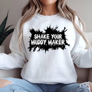 May include: White sweatshirt with the phrase "SHAKE YOUR MUDDY MAKER" in bold, black lettering. The text is set against a black, splattered design. The sweatshirt is a casual, comfortable style.