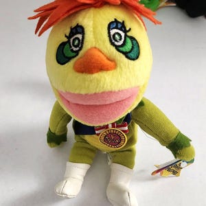 May include: A plush toy character with a yellow head, orange hair, and green eyes. The character wears a green outfit, white boots, and a medal that says "Mayor". A small tag is attached to the character's hand.
