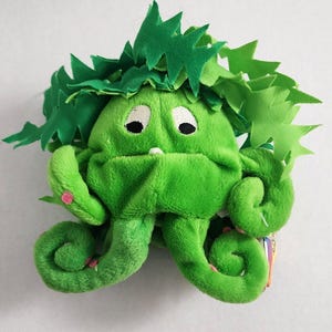 May include: A plush green monster toy with a leafy crown and curly tentacles. The monster has black eyes, a small white tooth, and pink accents on its arms. The toy is made of soft, fuzzy material.