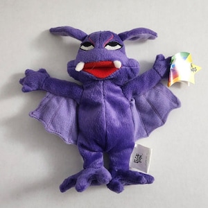 May include: A plush purple bat toy with outstretched wings, a red mouth, white fangs, and a stern expression. The toy has a tag attached. The bat's wings are a lighter shade of purple. The toy is a stuffed animal.