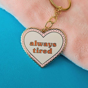 May include: A gold heart-shaped keychain with a white background and pink trim. The text "always tired" is printed in orange on the keychain.