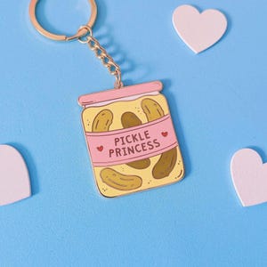May include: A gold-coloured keyring with a charm depicting a jar of pickles. The jar is yellow with brown pickles inside and a pink label reading "Pickle Princess" with heart accents. The background is light blue.