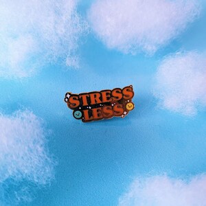 May include: A gold-coloured enamel pin with the words "Stress Less" in orange letters. The pin has two small smiley faces, one blue and one yellow.