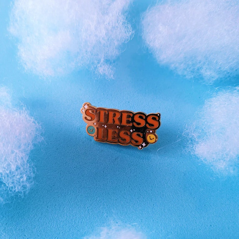 May include: A gold-coloured enamel pin with the words "Stress Less" in orange letters with a blue and yellow smiley face on each side of the text.