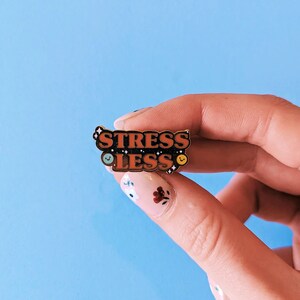 May include: A gold enamel pin with the words "Stress Less" in orange letters on a black background. The pin has two small smiley faces on either side of the text.