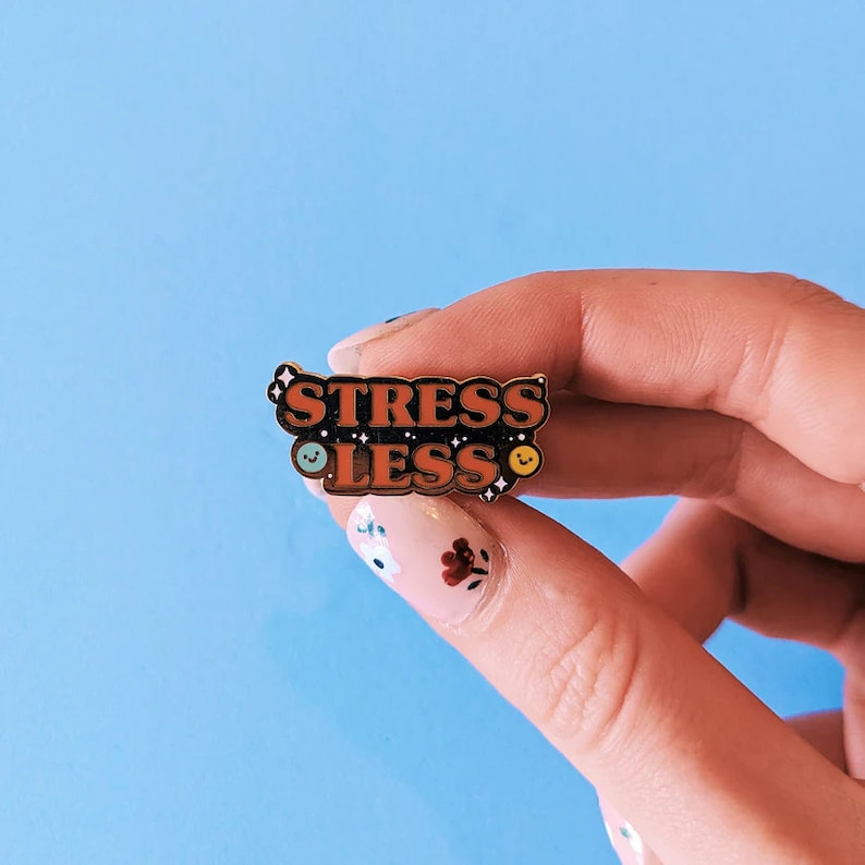 May include: A gold enamel pin with the words "Stress Less" in orange letters on a black background. The pin has two small smiley faces on either side of the text.