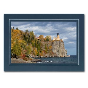 May include: A yellow lighthouse with a black roof sits on a rocky cliff overlooking a blue lake. The cliff is covered in trees with fall foliage in shades of red, orange, and yellow. The sky is cloudy and blue.
