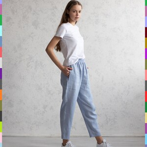 May include: A person wearing a white t-shirt and light blue linen pants. The pants have an elastic waistband and pockets. White sneakers complete the casual outfit. The background is a light gray textured wall.