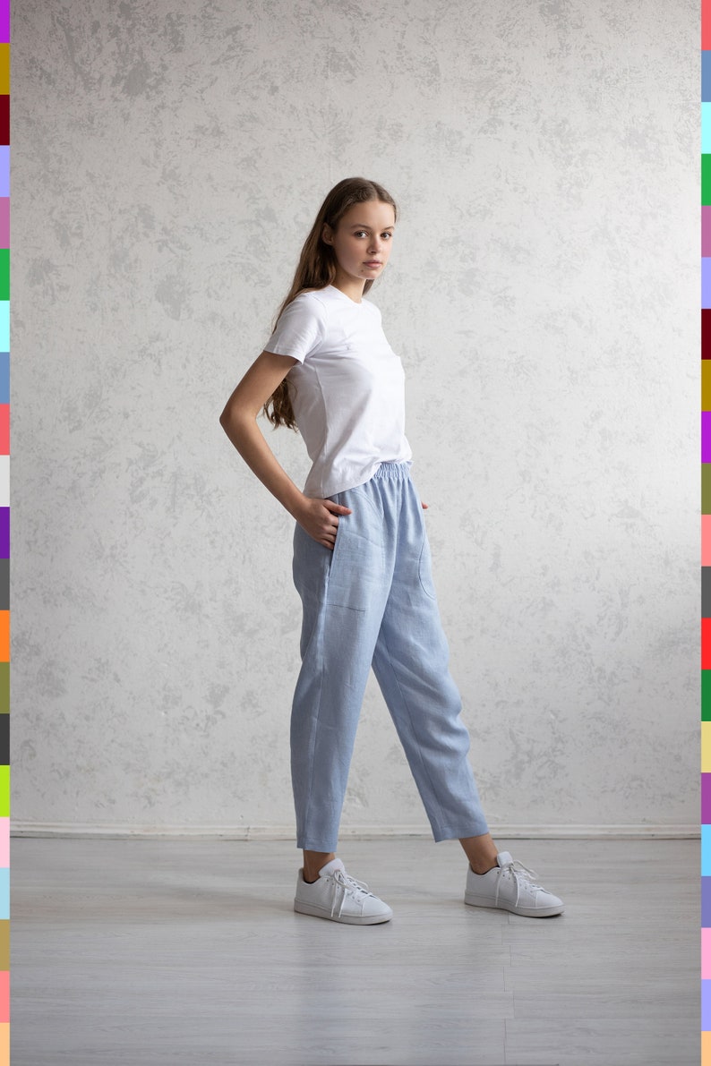 May include: A person wearing a white t-shirt and light blue linen pants. The pants have an elastic waistband and pockets. White sneakers complete the casual outfit. The background is a light gray textured wall.