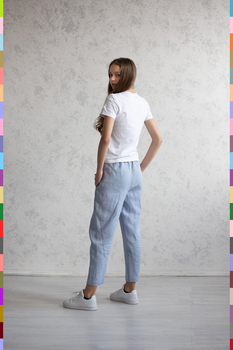 May include: A person wearing a white t-shirt and light blue linen pants. The pants have a relaxed fit and are cuffed at the ankles. White sneakers complete the outfit. The background is a textured gray wall.